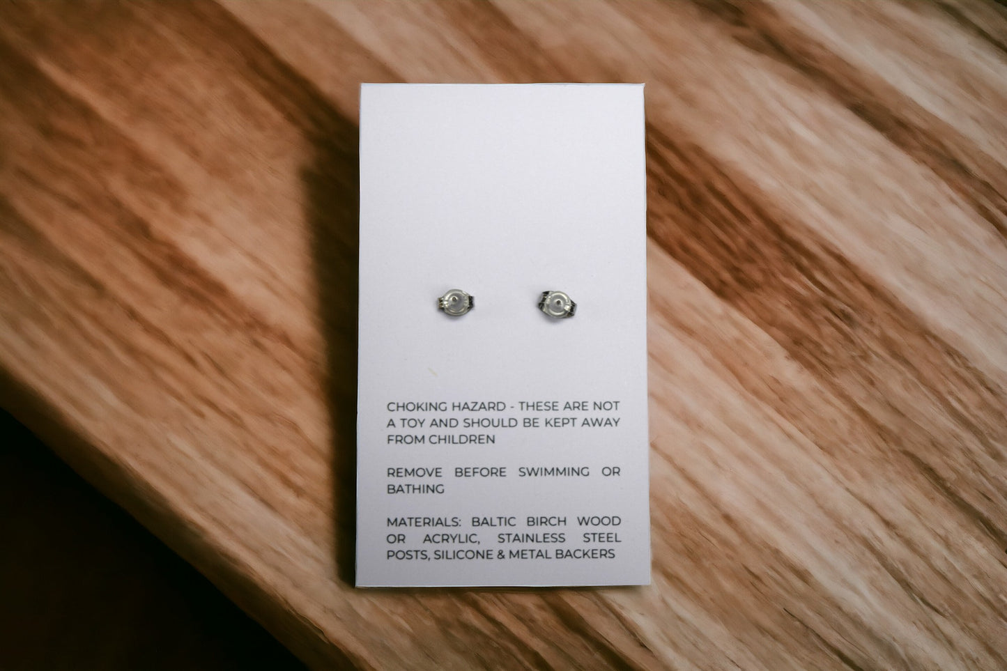 the back of an earring card showing posts and metal and silicone backers and care information for earrings laying flat on wood background