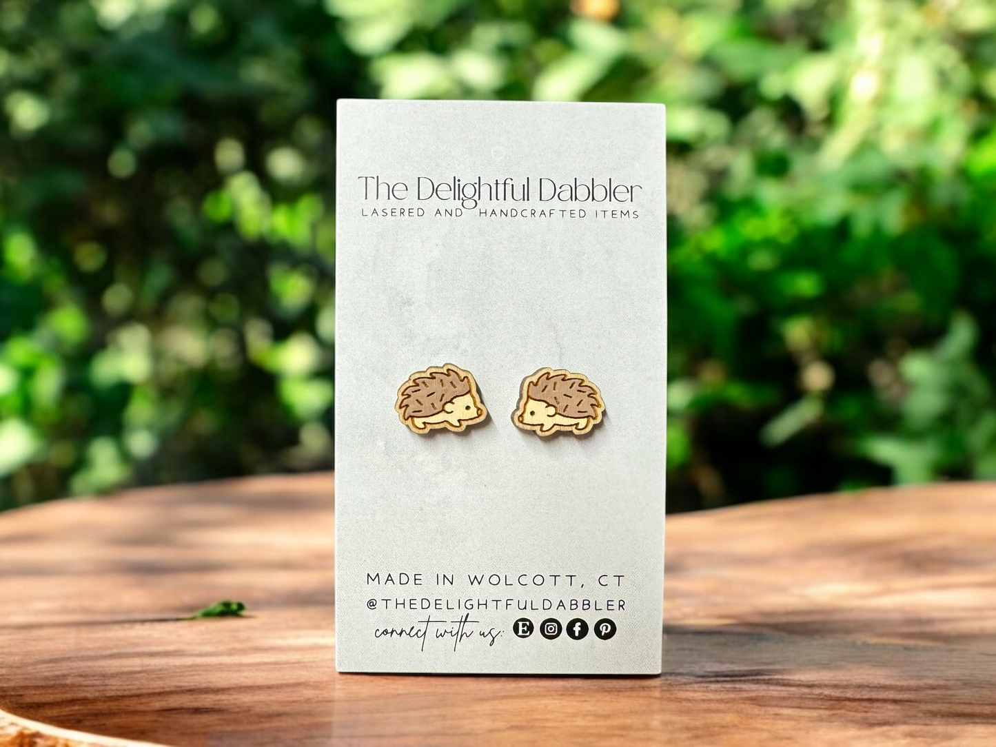 handpainted hedgehog-shaped wood stud earrings on a card with 'The Delightful Dabbler' branding, set against a blurred green outdoor background.