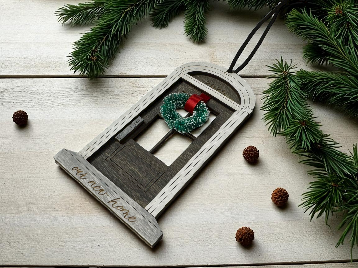 Decorative ornament shaped like a house with a wreath, on a wooden surface with pine branches and cones.