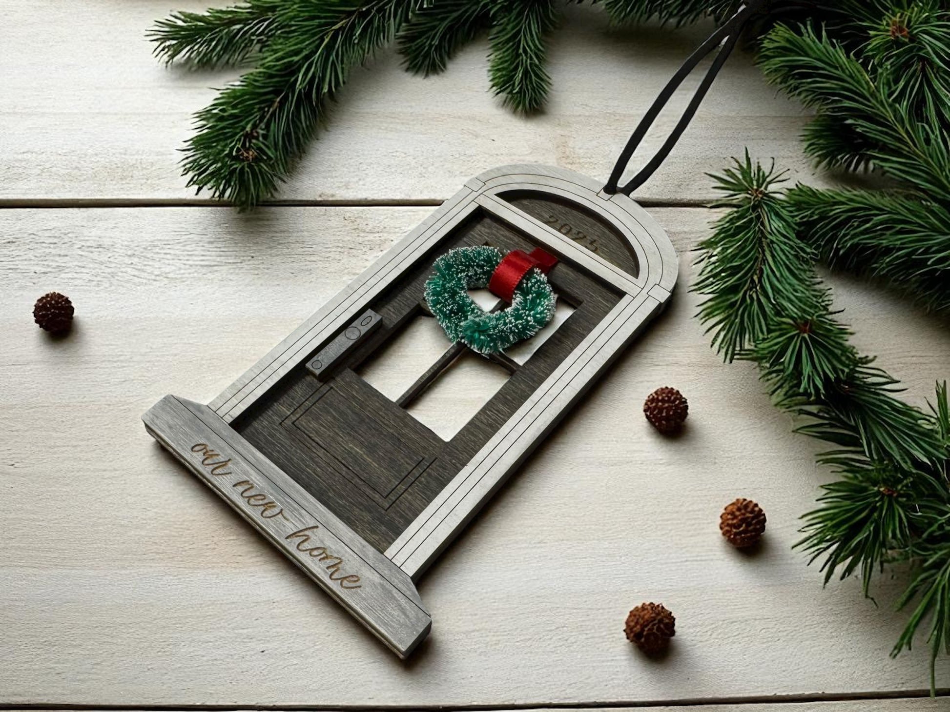 Decorative ornament shaped like a house with a wreath, on a wooden surface with pine branches and cones.