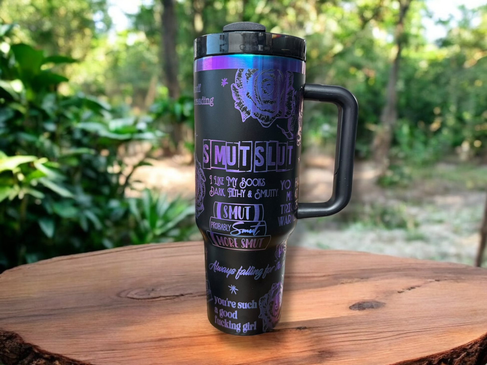 A 40oz black powder coated stainless steel tumbler with smutty images and sayings engraved all over exposing blue/purple metal, displayed on a wooden surface with a greenery background.