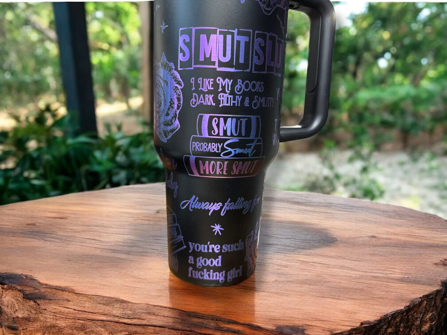 A 40oz black powder coated stainless steel tumbler with smutty images and sayings engraved all over exposing blue/purple metal, displayed on a wooden surface with a greenery background.