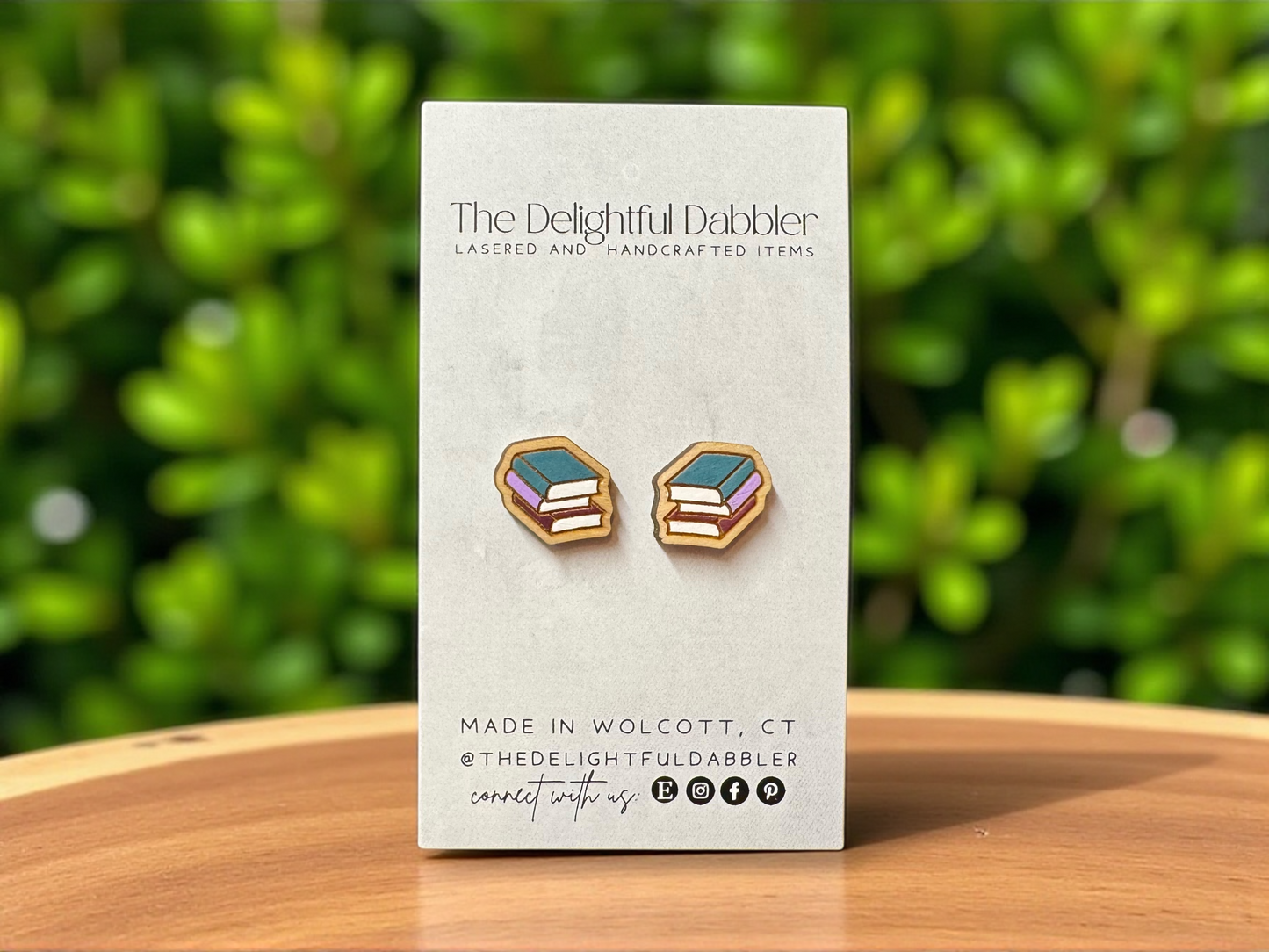 handpainted wood book stack stud Earrings on a card with 'The Delightful Dabbler' branding against a green leafy background.
